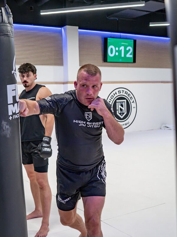 Frank squares up in gloves beside the heavy bags with a student behind him