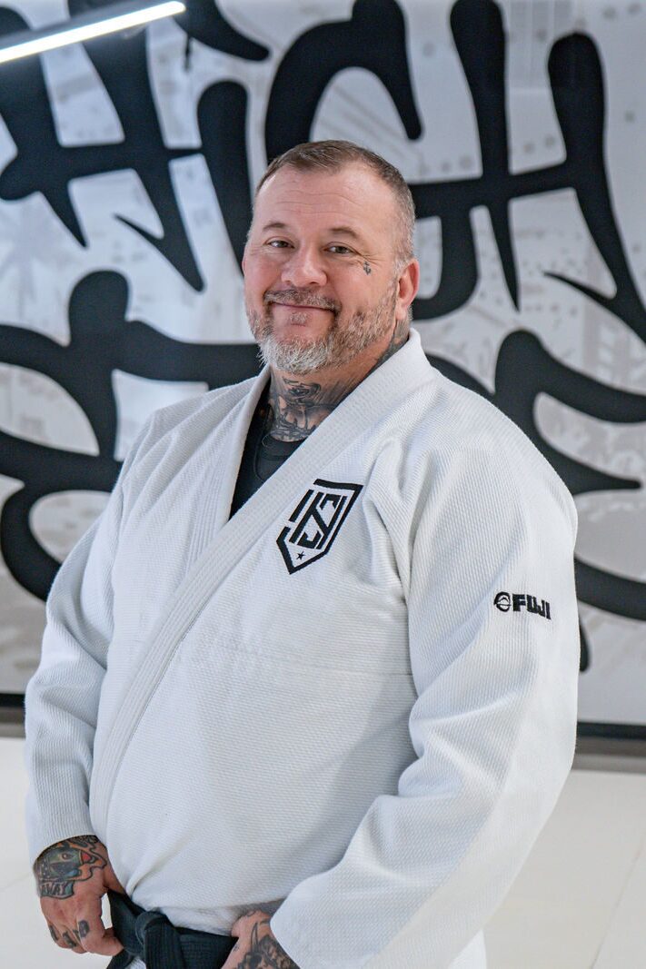 Coach Josh Halstead smiling in a white gi in front of the High Street mural