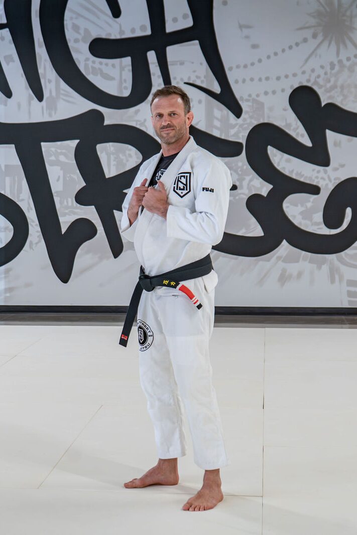 Coach Jay Shellhammer standing alone on the mats in a white gi and black belt, smiling, with the High Street mural behind him