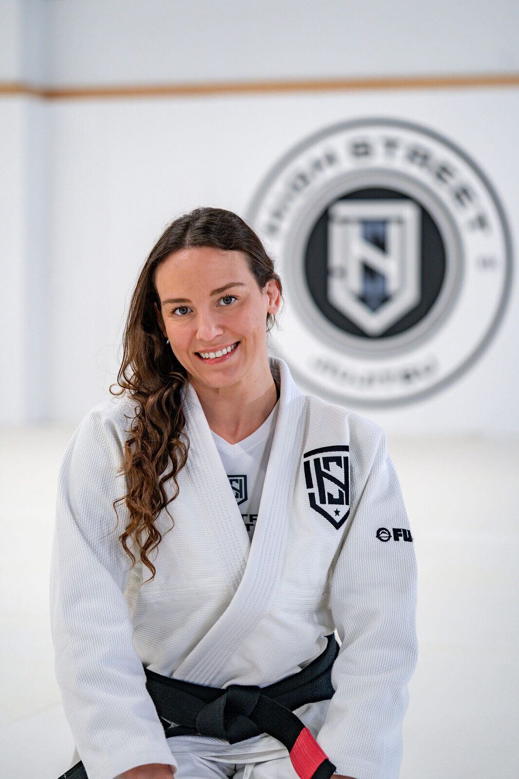 Coach Karla Shellhammer smiling in a white gi and black belt beneath the High Street seal