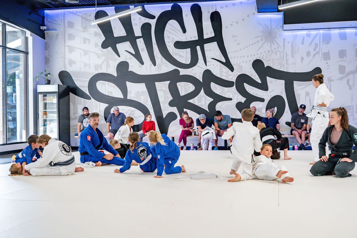 A kids class sits on the mat while parents watch from the benches