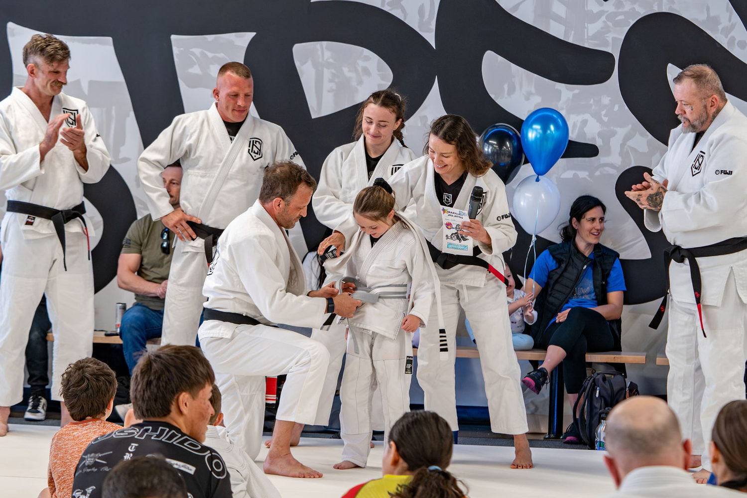 A coach ties on a young student's new belt while coaches and families celebrate