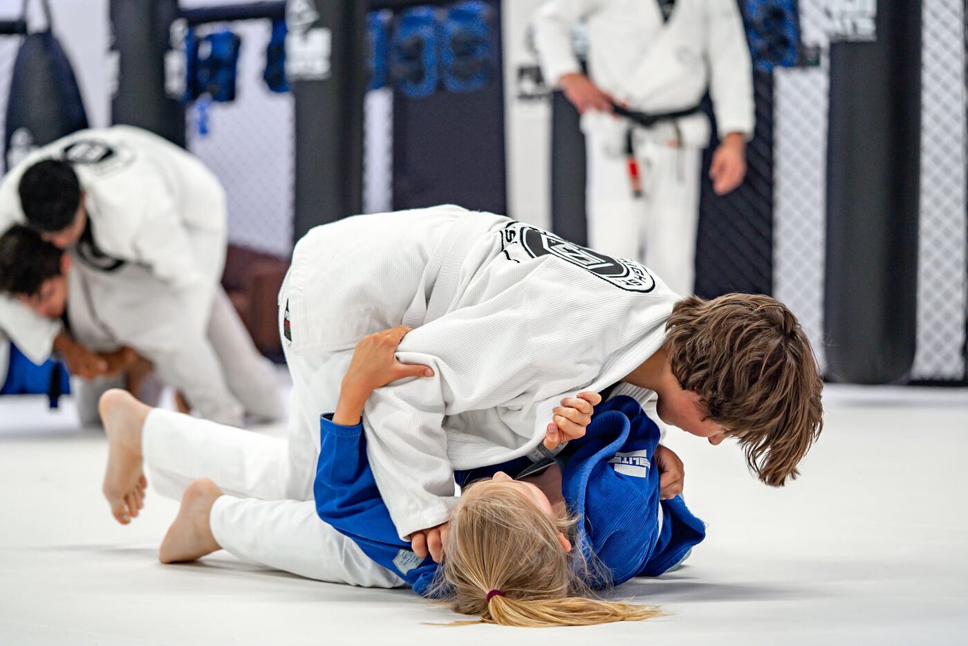 Two young students in white and blue gis roll on the mats during kids class, with a coach watching behind them