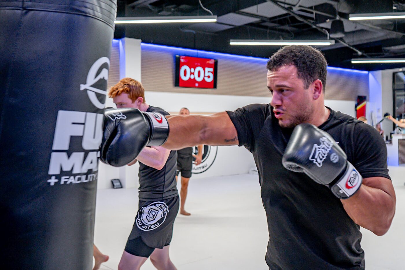 A striker in gloves lands a straight punch on the heavy bag under the round clock