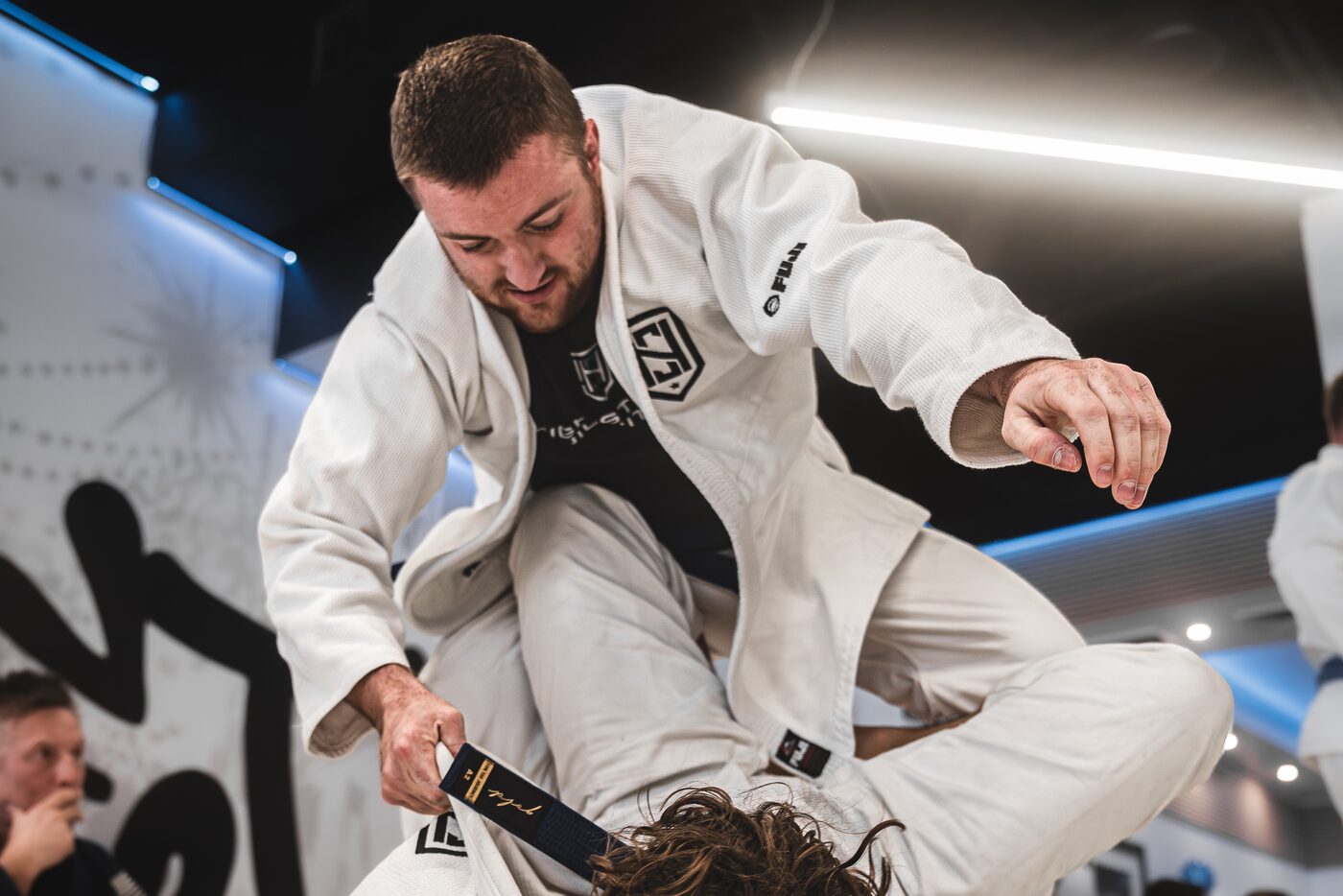 A coach in a white gi works a guard pass over a student on the mats while the class watches from the wall