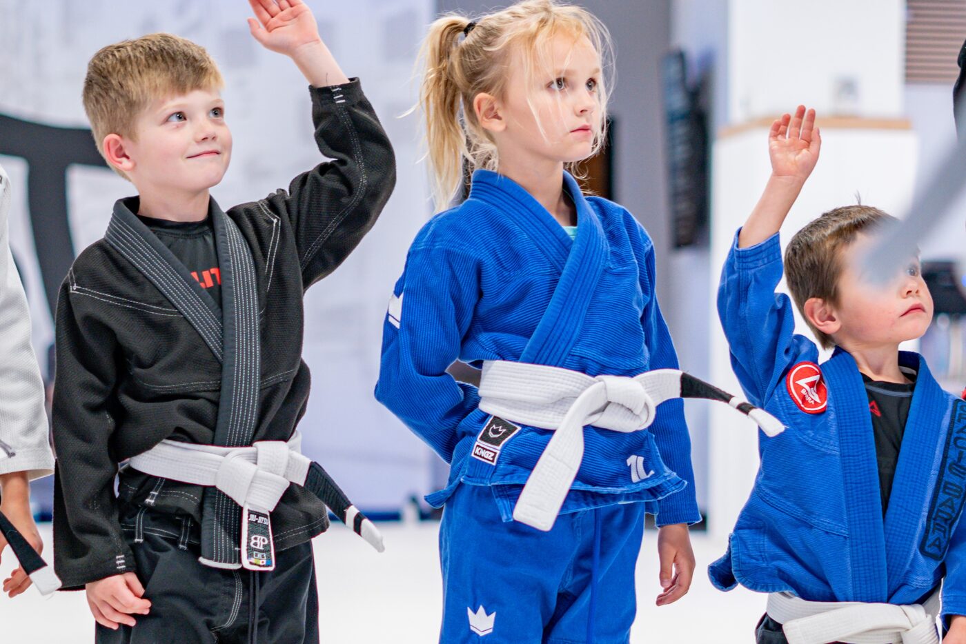 Three young students in gis stand in a line on the mats with their hands raised to answer their coach